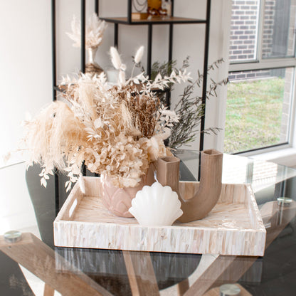 Nanaya 45cm Mother of Pearl Decorative Tray in Cream Beige features a vase with dried flowers, a U-shaped wooden holder, and a scallop-shaped object on a glass table.