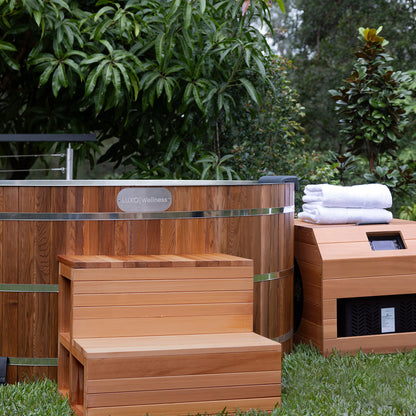 Denman 2 Person Outdoor Ice Bath with 1.5HP Chiller in Natural, features a barrel-style wooden tub on a lawn, accompanied by a matching two-step bench and equipment box with white towels.