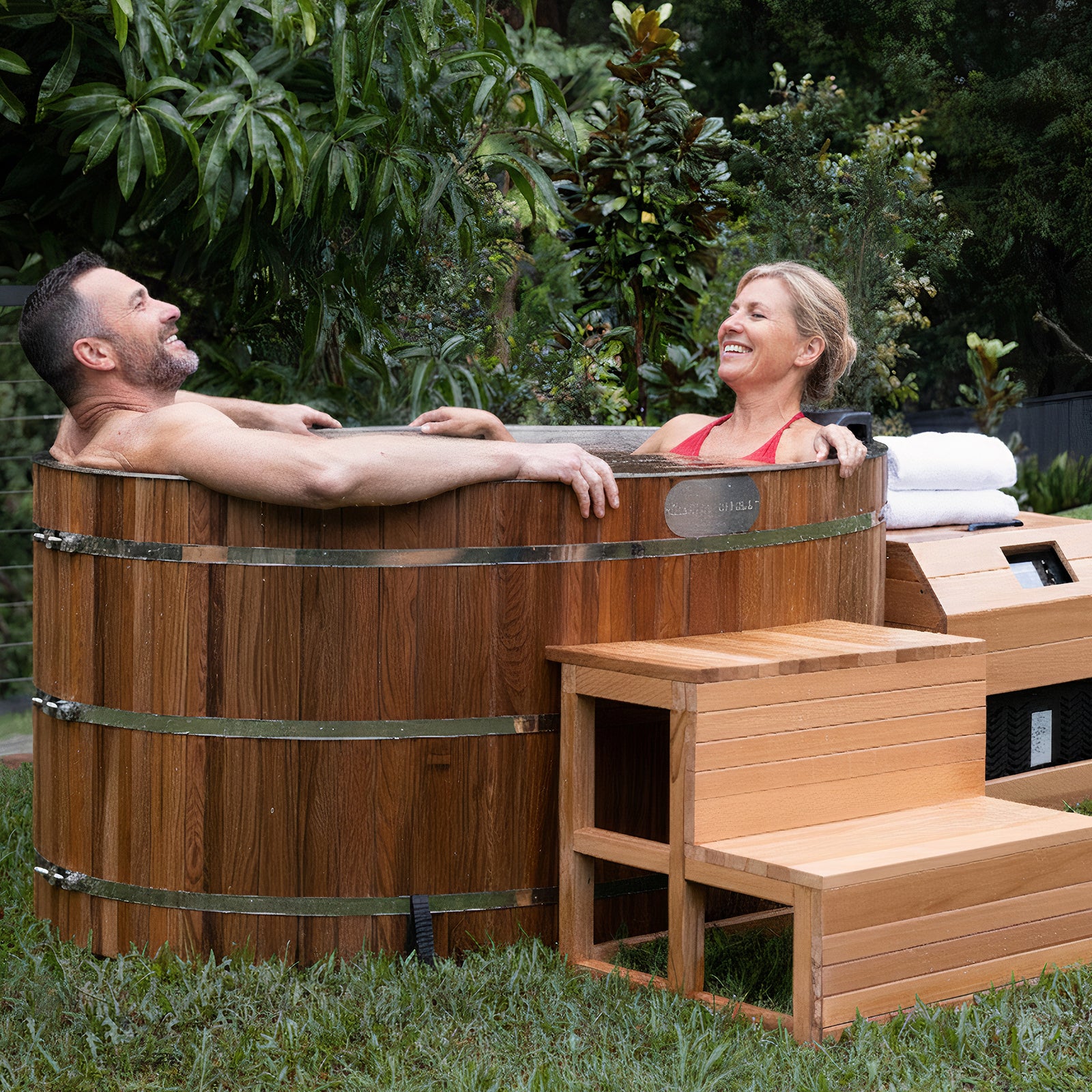 A couple relaxes in the Denman 2-Person Outdoor Ice Bath with 1.5HP Chiller, featuring a round wooden design with metal bands, set on a lawn with towels and a step stool nearby.