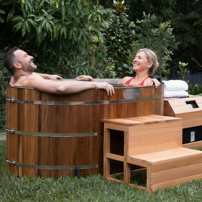 A couple relaxes in the Denman 2-Person Outdoor Ice Bath with 1.5HP Chiller, featuring a round wooden design with metal bands, set on a lawn with towels and a step stool nearby.