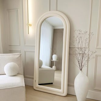 Auden 180cm Arch Full Length Mirror in Beige, with a textured off-white frame, leans against a cream wall beside a brass sconce, reflecting sheer curtains and a wooden slat wall.