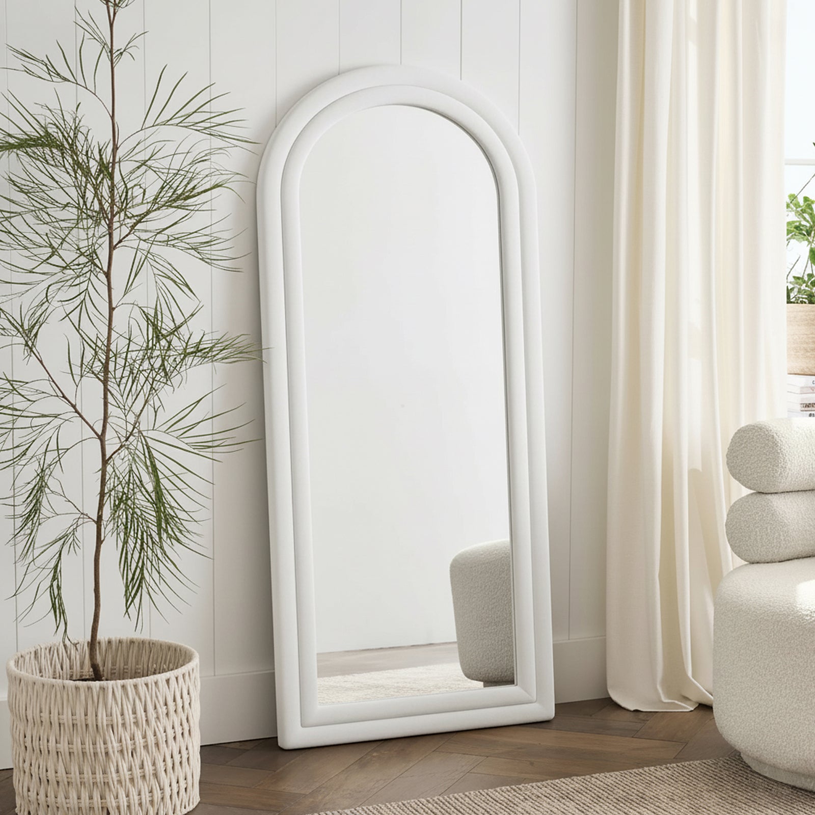 Auden 180cm Arch Full Length Mirror in Cream White, featuring a soft velvet frame, leans against a paneled wall, accompanied by a potted plant, boucle seat, and warm wood flooring.