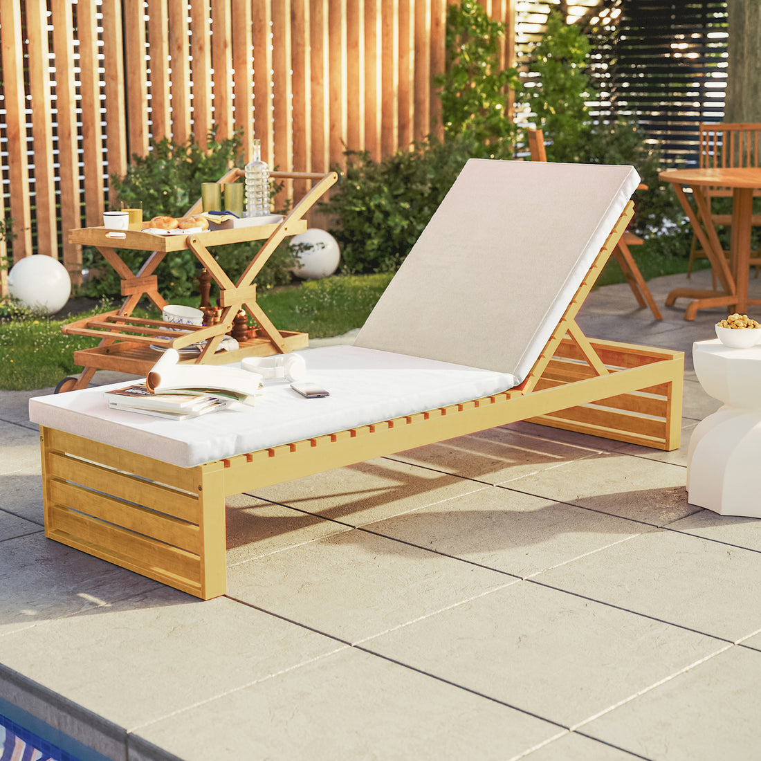 Alabat Acacia Wood Outdoor Sun Lounger with Cushion in Cream beside a pool, featuring an adjustable wooden frame, plush cushion, open magazine, smartphone, headphones, with a serving cart and side table nearby.