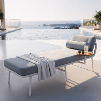 Andre Wicker Outdoor Sun Lounger with Cushion in Light Grey, featuring a modern chaise lounge with a matching throw, side table with iced drink, beside an infinity pool merging into a distant ocean.