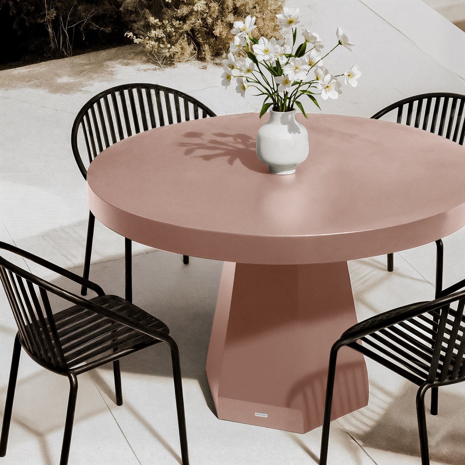 Apollo 120cm Outdoor Concrete Dining Table in Mocha Mousse with a terracotta-pink hue, round design, and sculptural pedestal base, surrounded by black metal chairs on a stone patio.