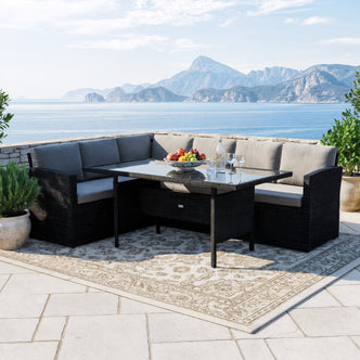 Ballena 6 Seater Black Wicker Outdoor Dining Set in Dark Grey on a seaside terrace with a glass-topped wicker table, light grey cushions, and surrounding potted plants, creating a serene coastal ambiance.