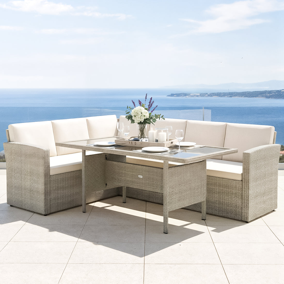 Ballena 6 Seater Warm Grey Wicker Outdoor Dining Set in Beige features an L-shaped sectional sofa with cushions, a rectangular dining table, a vase, candles, and place settings, overlooking the ocean.