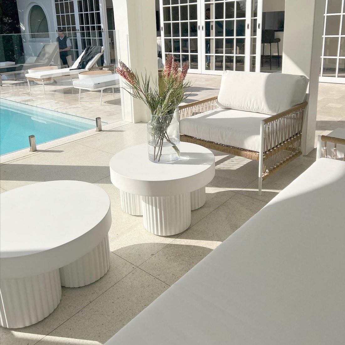 Basalt 70cm Indoor & Outdoor Concrete Coffee Table in White, featuring a round top and fluted base, adorned with a glass vase of pink flowers, set by a sunlit poolside.