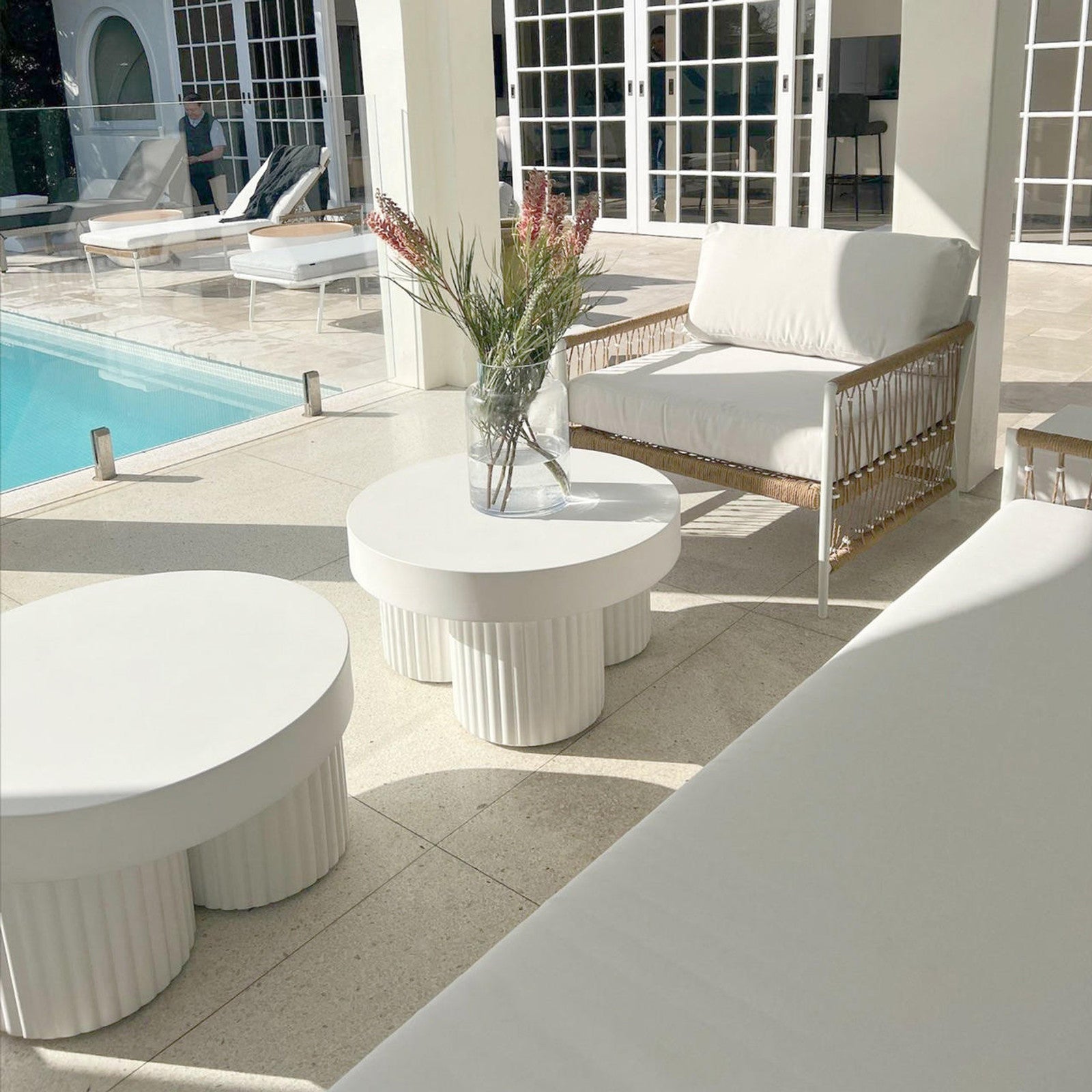 Basalt 70cm Indoor & Outdoor Concrete Coffee Table in White, featuring a round top and fluted base, adorned with a glass vase of pink flowers, set by a sunlit poolside.
