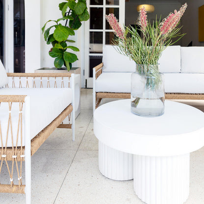 Basalt 70cm Indoor & Outdoor Concrete Coffee Table in White, featuring a sleek, minimalist design with a round shape, topped with a glass vase of pinkish flowers, set in a bright seating area.