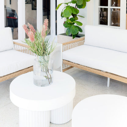Basalt 70cm Indoor & Outdoor Concrete Coffee Table in White, showcasing a sleek, minimalist design with a smooth surface, paired with a glass vase holding pinkish flowers, creating an elegant lounge atmosphere.