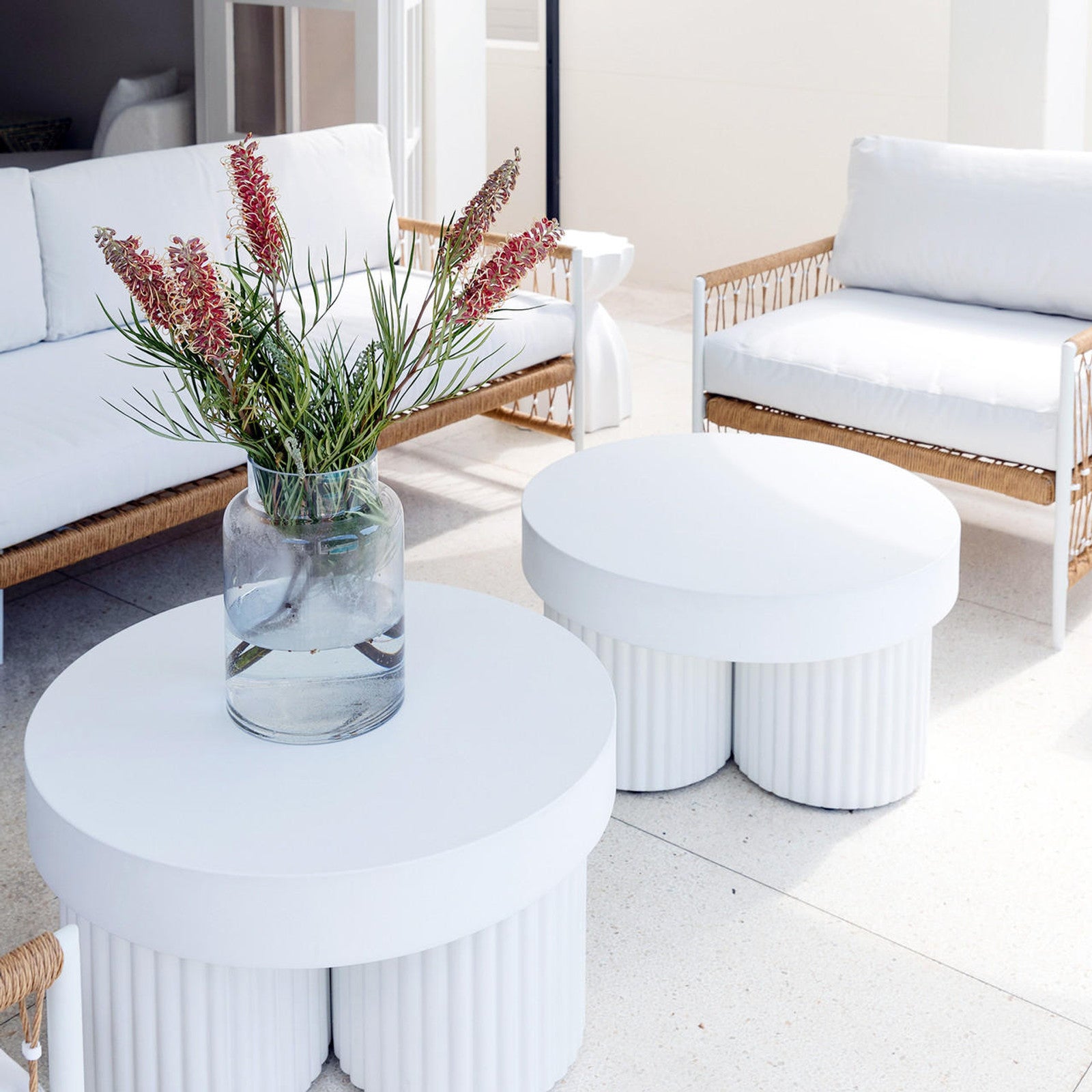 White Basalt 70cm Indoor/Outdoor Concrete Coffee Table with fluted cylindrical supports, set in a minimalist seating area with rattan loveseat, armchair, and spiky green-stemmed blooms in a vase.