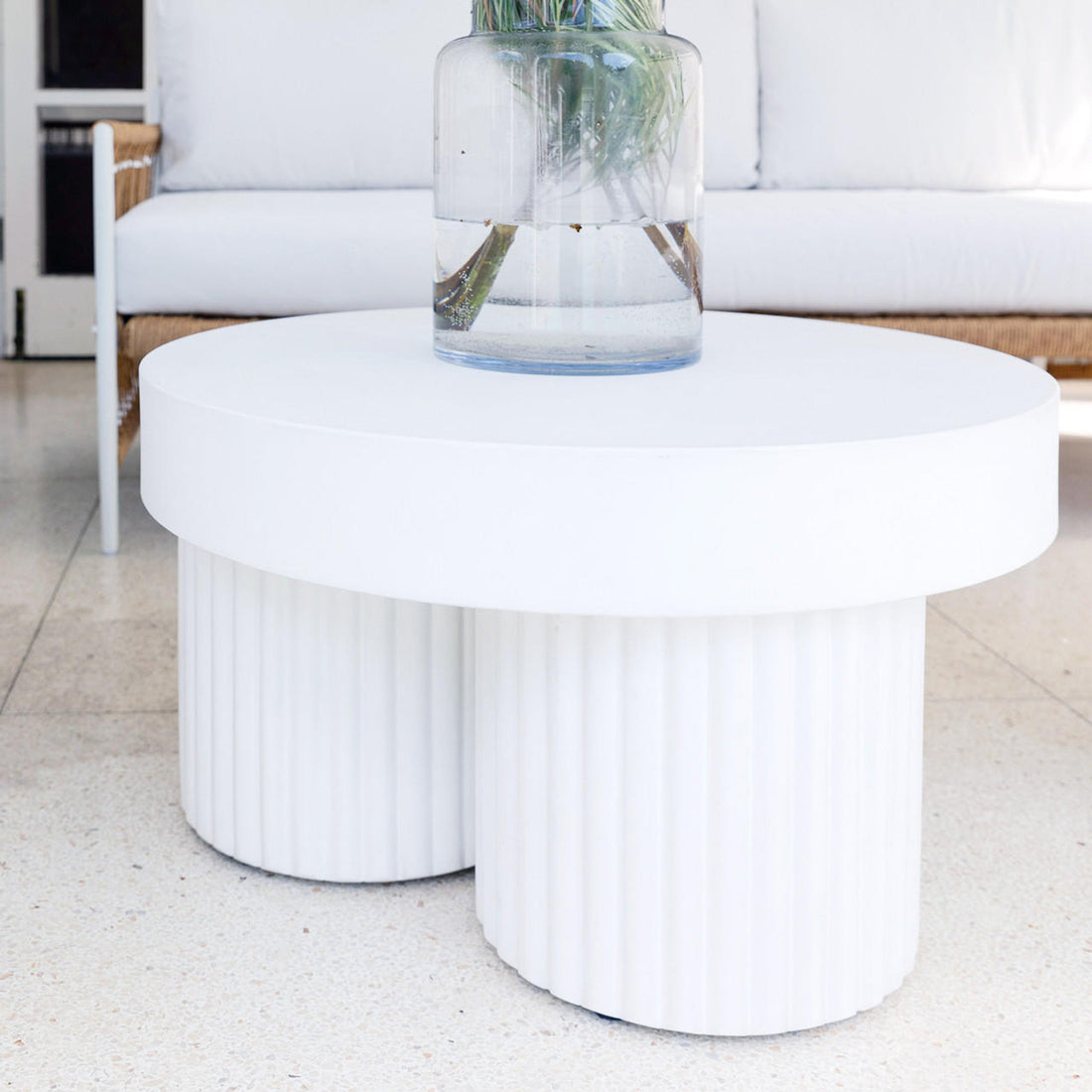 Basalt 70cm Indoor & Outdoor Concrete Coffee Table in White, featuring a round white top supported by fluted cylindrical bases, shown with a clear vase and green foliage on a terrazzo floor.
