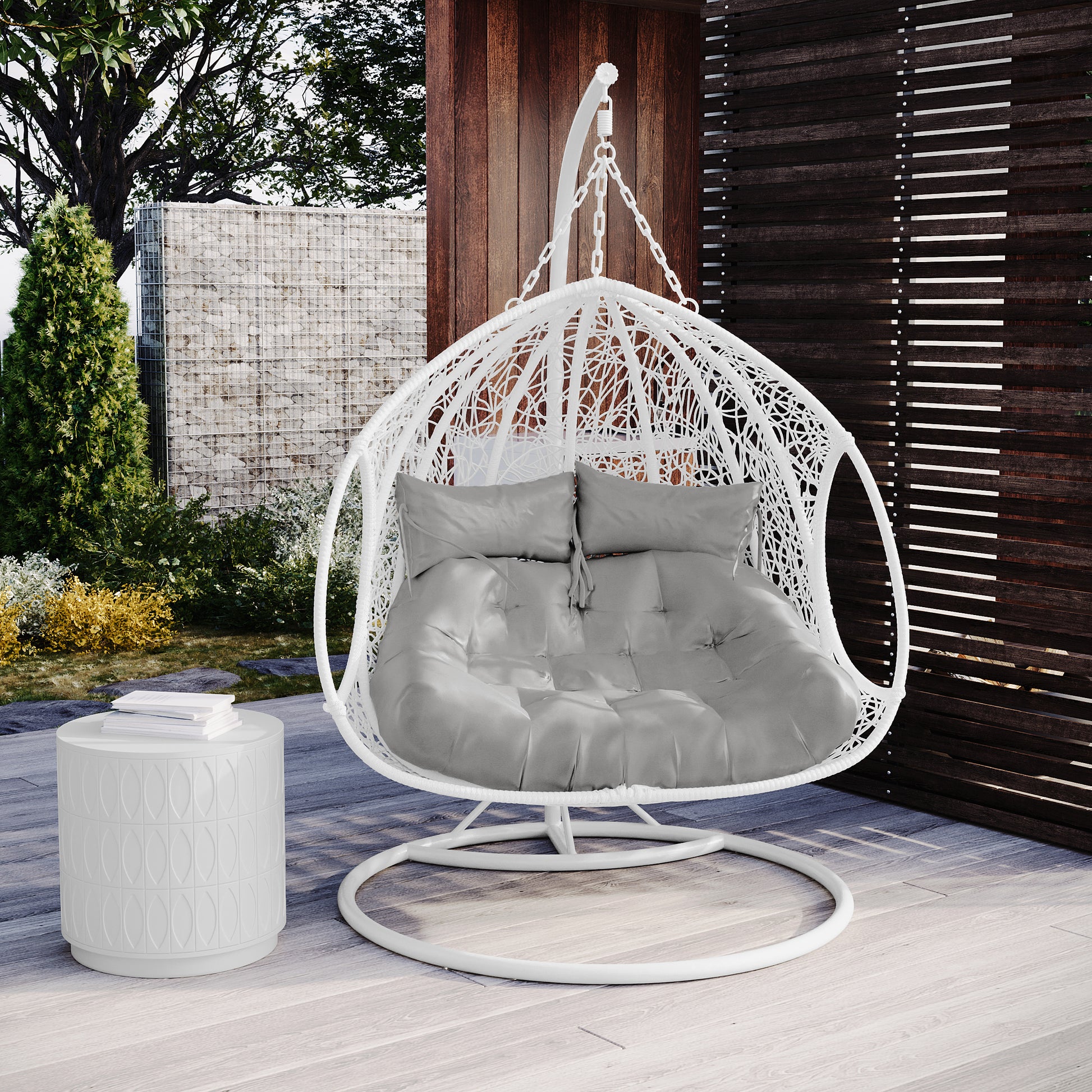 Basin 2-Seater Hanging Egg Chair in White, featuring a woven design, grey tufted cushions, and a matching frame, set on light wood decking with a privacy screen backdrop.