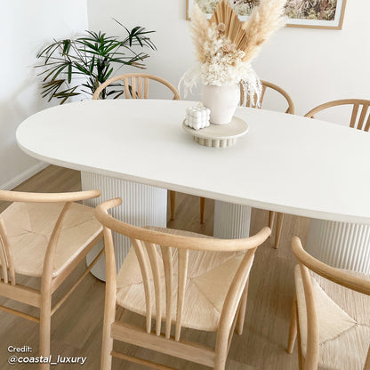 Belmar 200cm Concrete Indoor/Outdoor Dining Table in White, featuring an oval top with fluted pedestal legs, surrounded by Scandinavian-style chairs, styled with a vase of dried botanicals and a bubble candle.