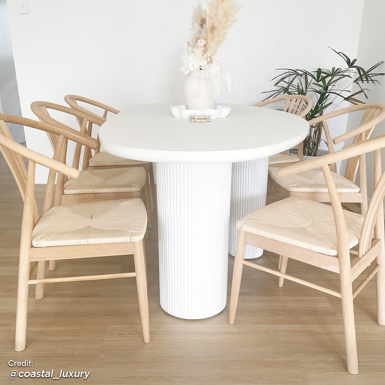 Belmar 200cm Concrete Dining Table in White with a round design, fluted cylindrical base, set with natural-wood chairs, dried floral centerpiece, and gold accents in a coastal-style dining nook.