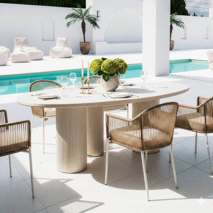 Belmar 200cm Curved Outdoor Concrete Dining Table in White with a fluted cylindrical base, surrounded by four woven-rope chairs, set with plates and a vase of hydrangeas, beside a pool.