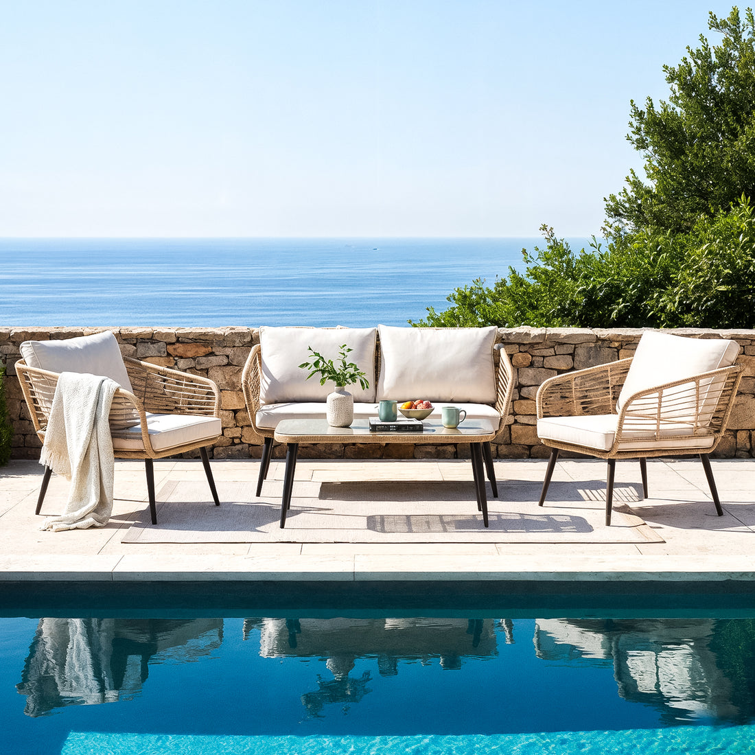Bequia 4-Seater Rope Wicker Outdoor Sofa in Sand, featuring a loveseat and two armchairs with cushions, set around a glass-topped coffee table on a sunlit poolside terrace overlooking the sea.