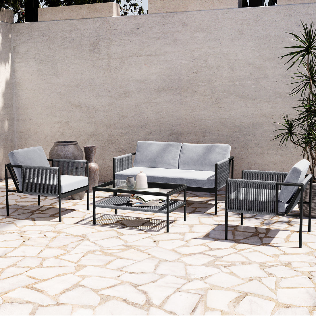 Brujas 4 Seater Wicker Outdoor Lounge Set in Grey, featuring a two-seat sofa, two armchairs, and a glass-top coffee table, elegantly arranged on a sunlit patio with decorative vases and greenery.