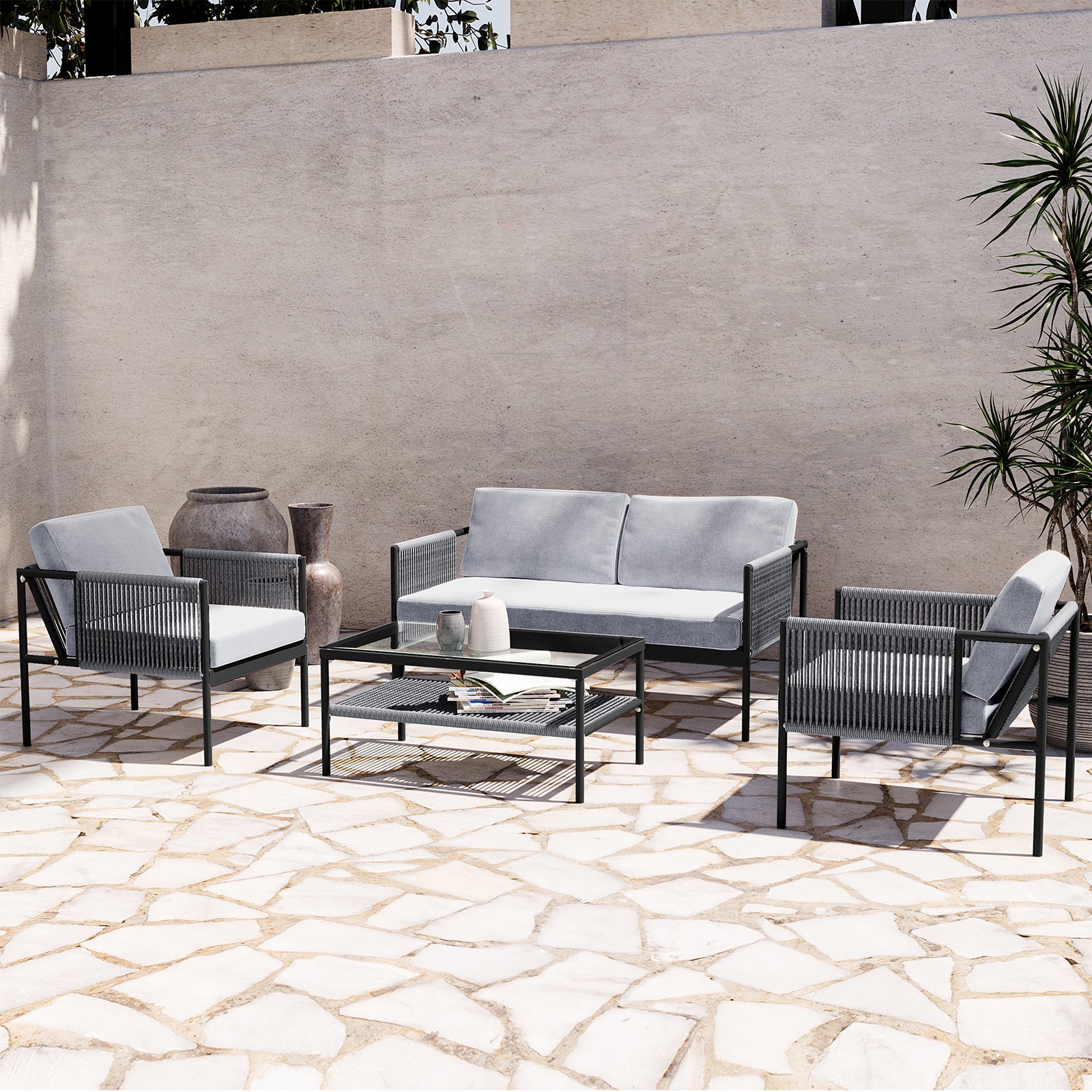 Brujas 4 Seater Wicker Outdoor Lounge Set in Grey, featuring a two-seat sofa, two armchairs, and a glass-top coffee table, elegantly arranged on a sunlit patio with decorative vases and greenery.
