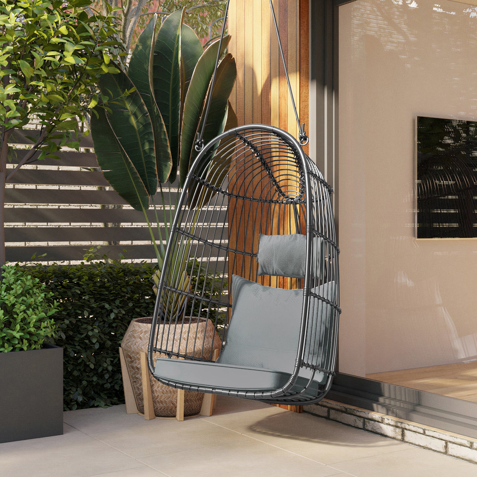 Burias Rattan Basket Outdoor Egg Chair with Rope in Black, featuring a suspended cocoon design with grey cushions, set on a patio with potted plants and a wooden privacy fence.
