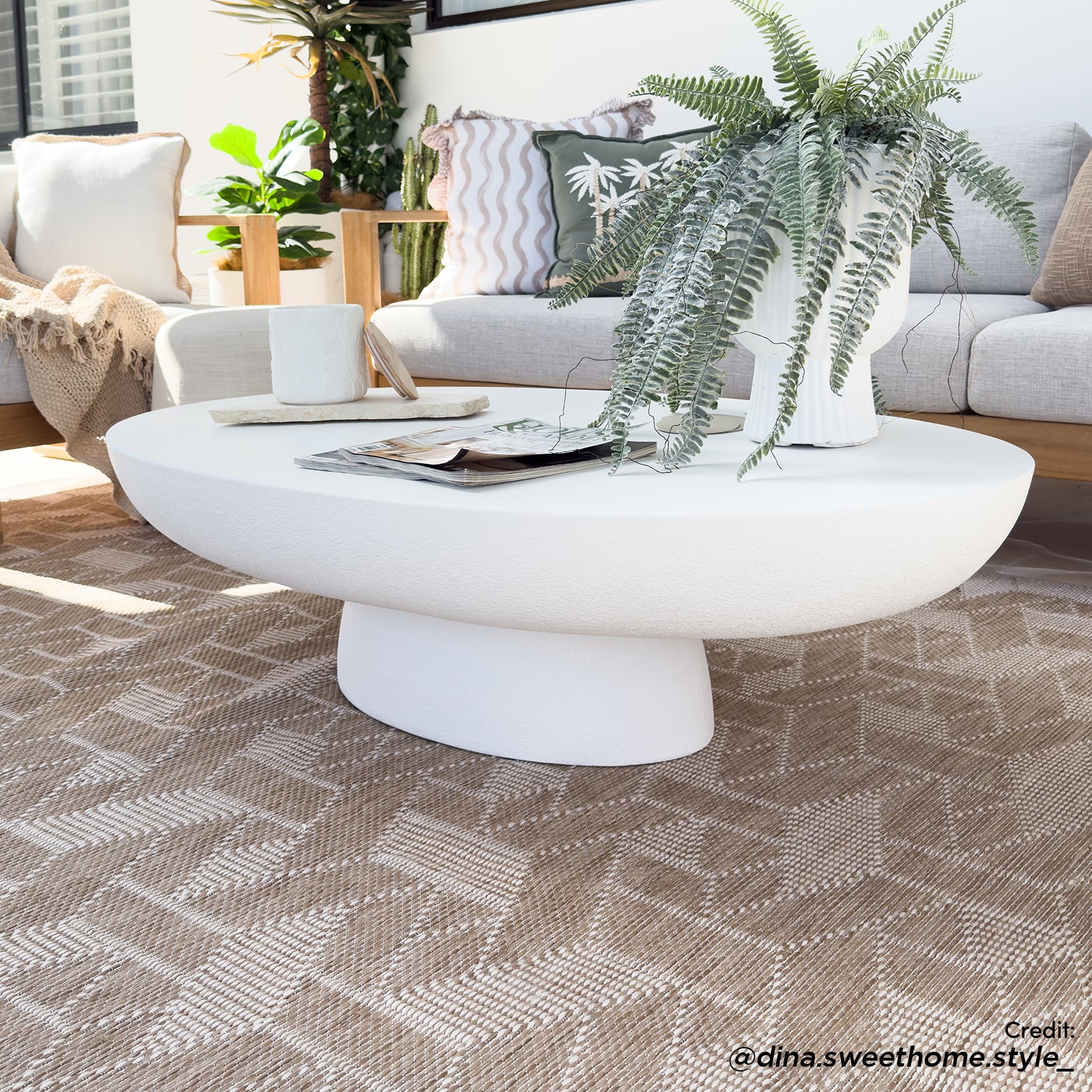 Cassius Oval Outdoor Concrete Coffee Table in White, featuring a smooth oval top and sculptural base, adorned with a lush fern, magazines, and a coaster, set on a geometric-patterned rug.