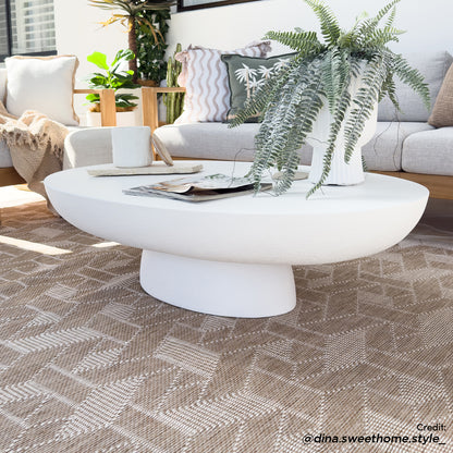 Cassius Oval Outdoor Concrete Coffee Table in White, featuring a smooth oval top and sculptural base, adorned with a lush fern, magazines, and a coaster, set on a geometric-patterned rug.