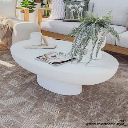 Cassius Oval Outdoor Concrete Coffee Table in White, central in a modern living room, features a sleek oval design. It's adorned with a lush fern, candle on tray, and magazines, exuding elegance.