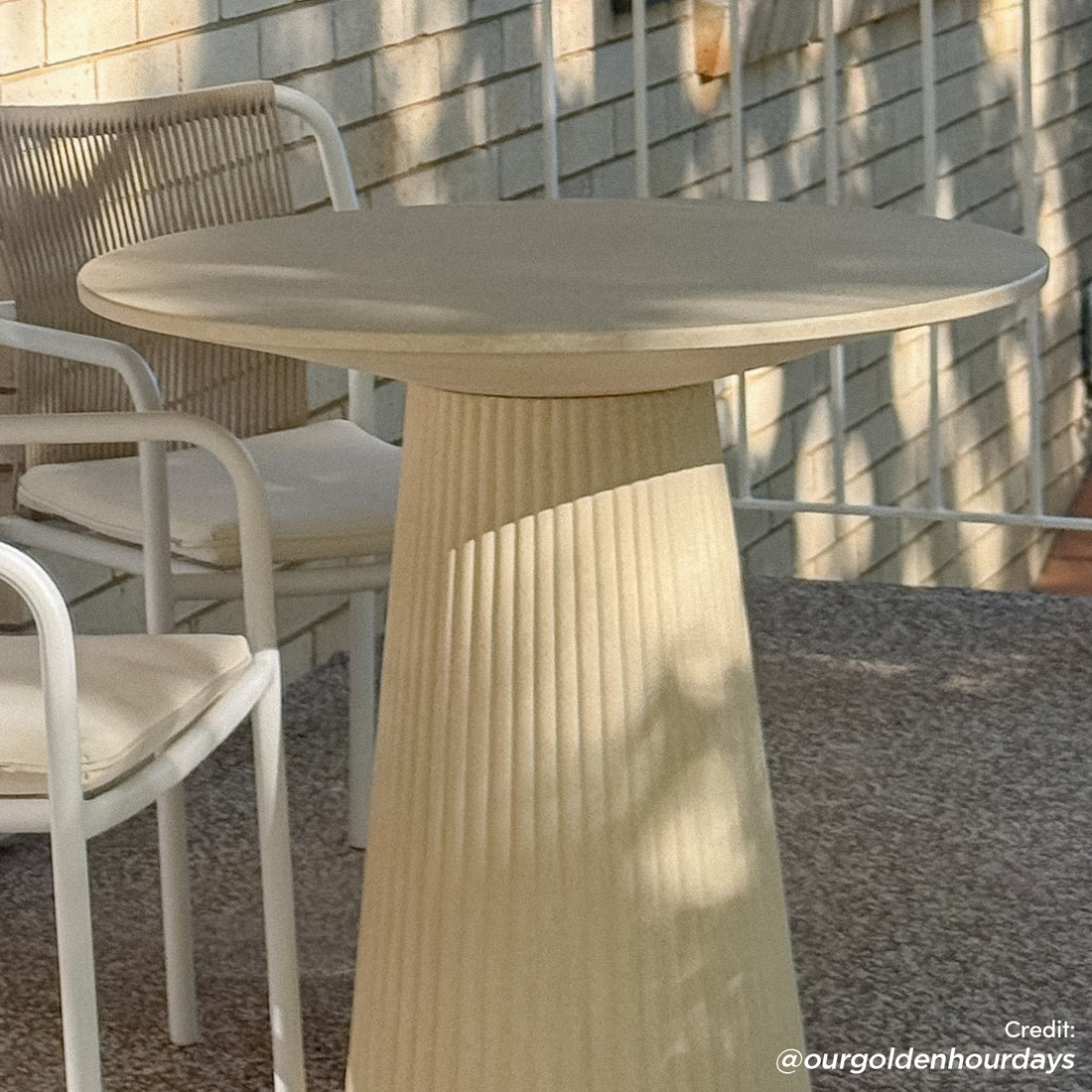 The Darien 80cm Round Concrete Bistro Table in Ivory features a fluted base and smooth top, styled with white-framed chairs on a textured outdoor surface against a pale brick backdrop.