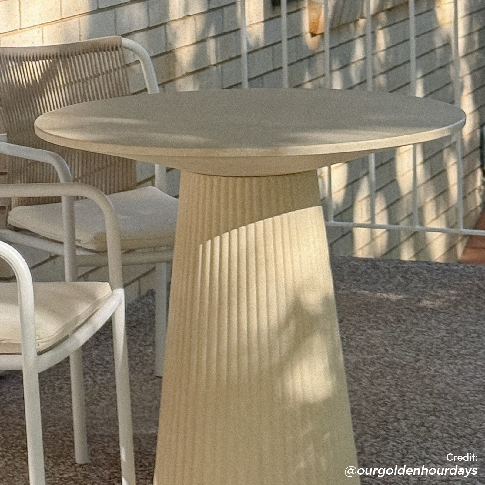 The Darien 80cm Round Concrete Bistro Table in Ivory features a fluted base and smooth top, styled with white-framed chairs on a textured outdoor surface against a pale brick backdrop.