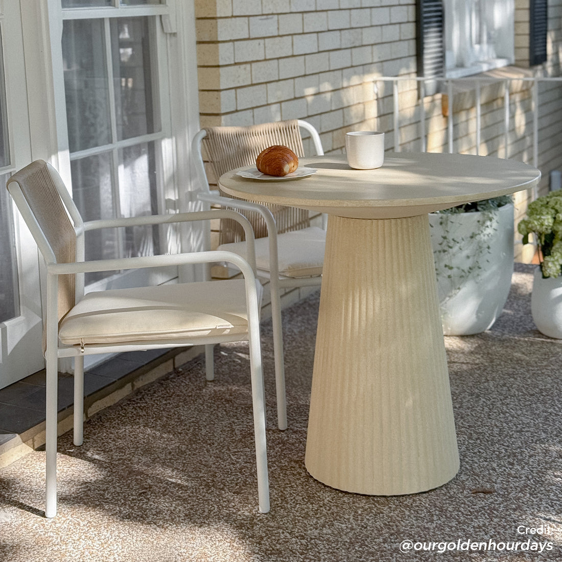 Darien 80cm Round Concrete Bistro Table in Ivory with a fluted pedestal base, set with a mug and croissant, flanked by two white-framed chairs with woven backs.