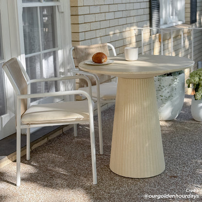 Darien 80cm Round Concrete Bistro Table in Ivory with a fluted pedestal base, set with a mug and croissant, flanked by two white-framed chairs with woven backs.