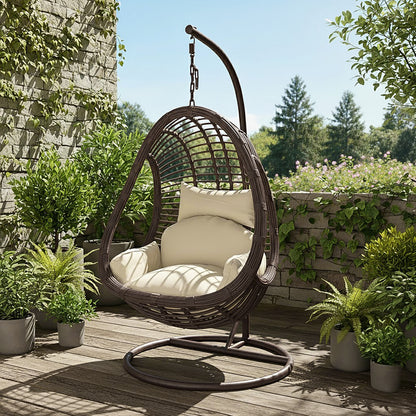 Deanna Curved Rattan Hanging Outdoor Egg Chair in Brown on a wooden deck, featuring a woven frame with cream cushions, surrounded by potted plants and vines against a stone wall.