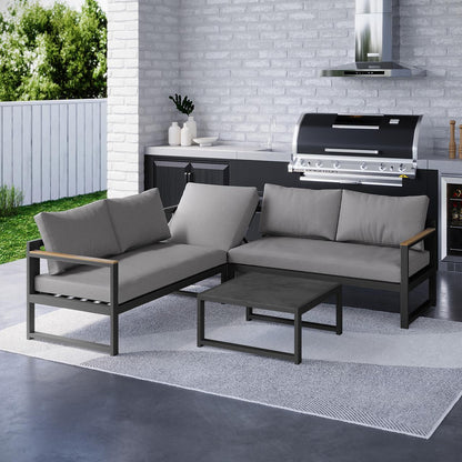 The Deloran 5-Seater Modular Corner Aluminium Outdoor Lounge Set Grey features an L-shaped sectional with gray cushions, paired with a matching coffee table, set against a white-washed brick wall patio.