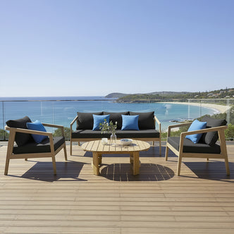 Delva 4 Seater Hardwood Acacia Outdoor Sofa Set in Black, featuring a two-seat sofa, two armchairs with charcoal cushions, and a round coffee table, set on a sunny coastal deck.
