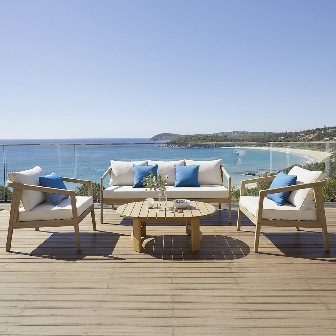 Delva 4 Seater Hardwood Acacia Outdoor Sofa Set in Cream on a seaside deck, featuring armchairs and sofa with cushions and blue throw pillows around a wooden coffee table, overlooking the beach.