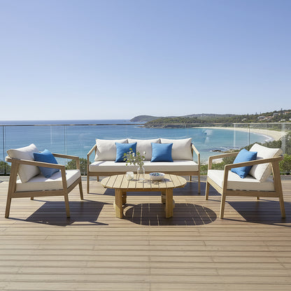 Delva 4 Seater Hardwood Acacia Outdoor Sofa Set in Cream on a seaside deck, featuring armchairs and sofa with cushions and blue throw pillows around a wooden coffee table, overlooking the beach.