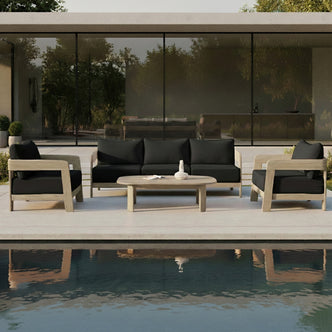 Elani 5 Seater Acacia Hardwood Outdoor Sofa Set in Black features a wooden-frame sofa and armchairs with black cushions, arranged around an oval coffee table on a sleek concrete terrace.