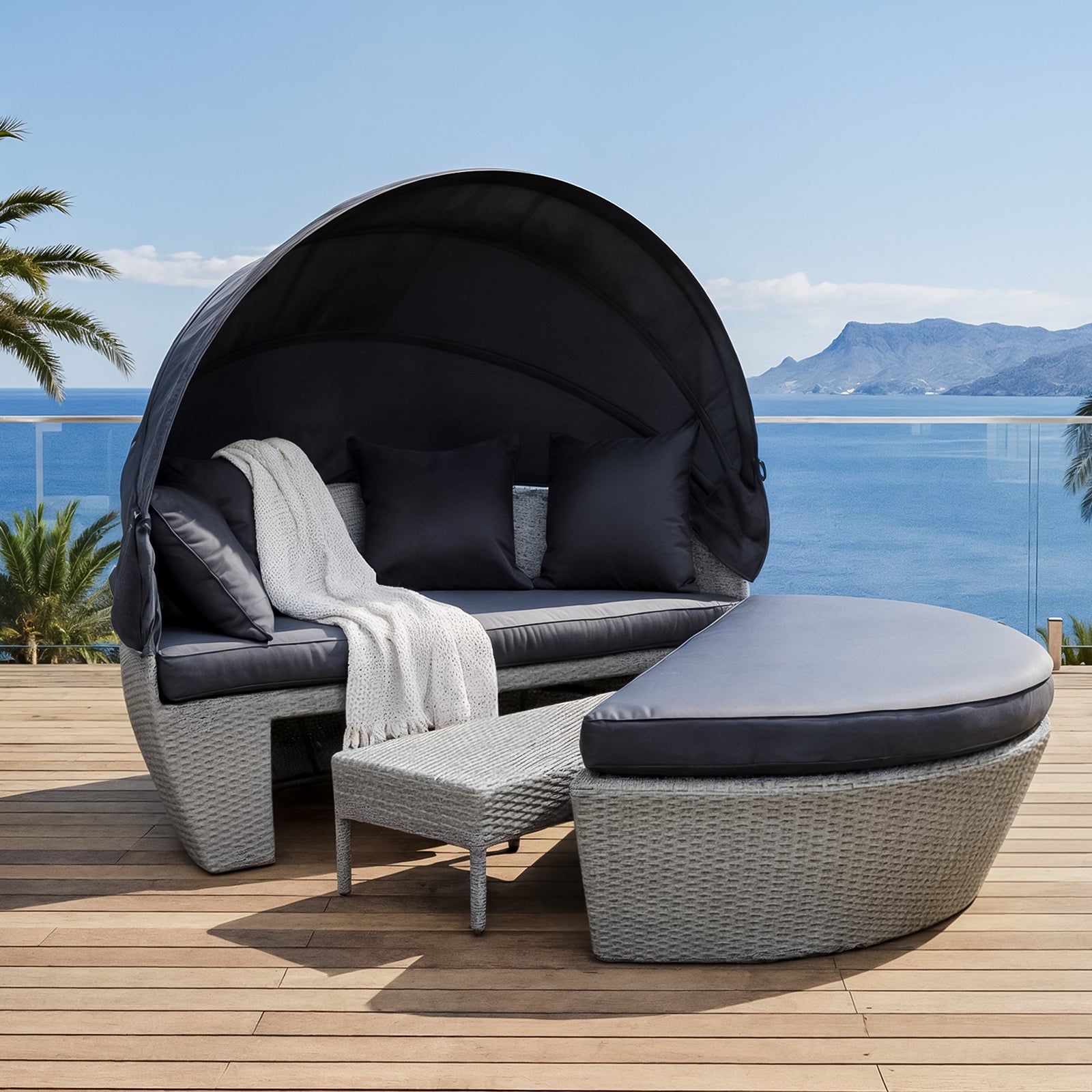 Erith Round Wicker Outdoor Canopy Daybed in Brown & Black featuring a semi-circular wicker design, retractable canopy, dark cushions, throw pillows, and an ottoman, set on a wooden deck overlooking the sea.