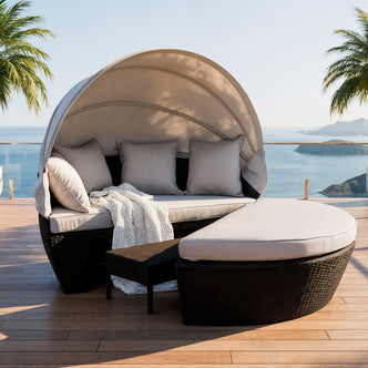 Erith Round Wicker Outdoor Daybed in Light Grey & Black features a semi-circular wicker design with a canopy, light-beige cushions, throw pillows, and a matching ottoman on a seaside deck.