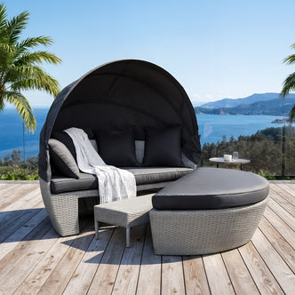 Erith Wicker Outdoor Canopy Daybed in Charcoal & Grey on a deck, featuring a woven design, curved canopy, dark cushions, a semi-circular ottoman, and a round side table with a glass.