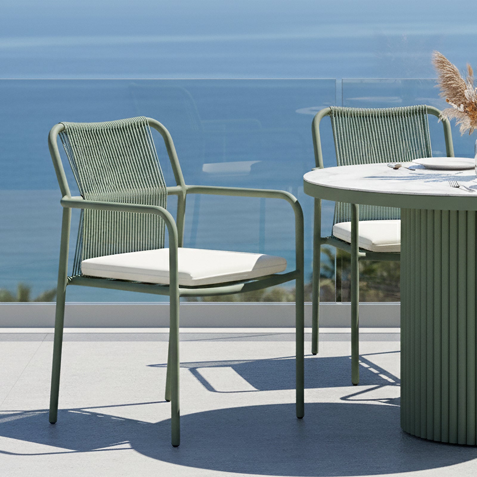Evone Aluminium Rope Outdoor Dining Chairs Set of 4 in Green, featuring metal frames and woven rope backs with white cushions, positioned on a seaside terrace with a round dining table nearby.