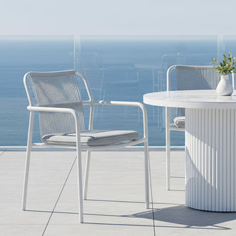 Evone Aluminium Rope Outdoor Dining Chairs Set of 4 in Light Grey on a terrace, featuring white frames, cord backrests, light gray cushions, and a round white table with green foliage against an ocean backdrop.