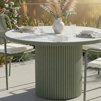 Evone Aluminium Outdoor Dining Table in Green with a round, fluted base, white stone-look top, set with simple place settings, and a vase of dried grasses, flanked by matching chairs.