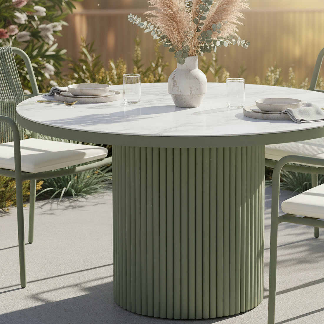 Evone Aluminium Outdoor Dining Table in Green with a round, fluted base, white stone-look top, set with simple place settings, and a vase of dried grasses, flanked by matching chairs.