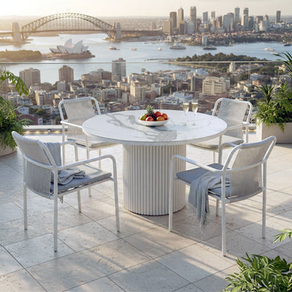 The Evone 4 Seater Stone Outdoor Dining Table Set in Light Grey features a round, marble-look tabletop with a white pedestal base, surrounded by modern rope-back chairs on a sunlit balcony.

