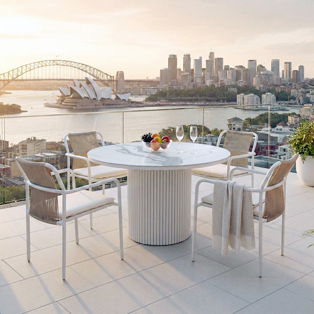Evone 4 Seater Stone Outdoor Dining Table Set featuring a round marble top with four rope-back chairs, glass bowl of fruit, and wine glasses, overlooking a harbor at sunset.