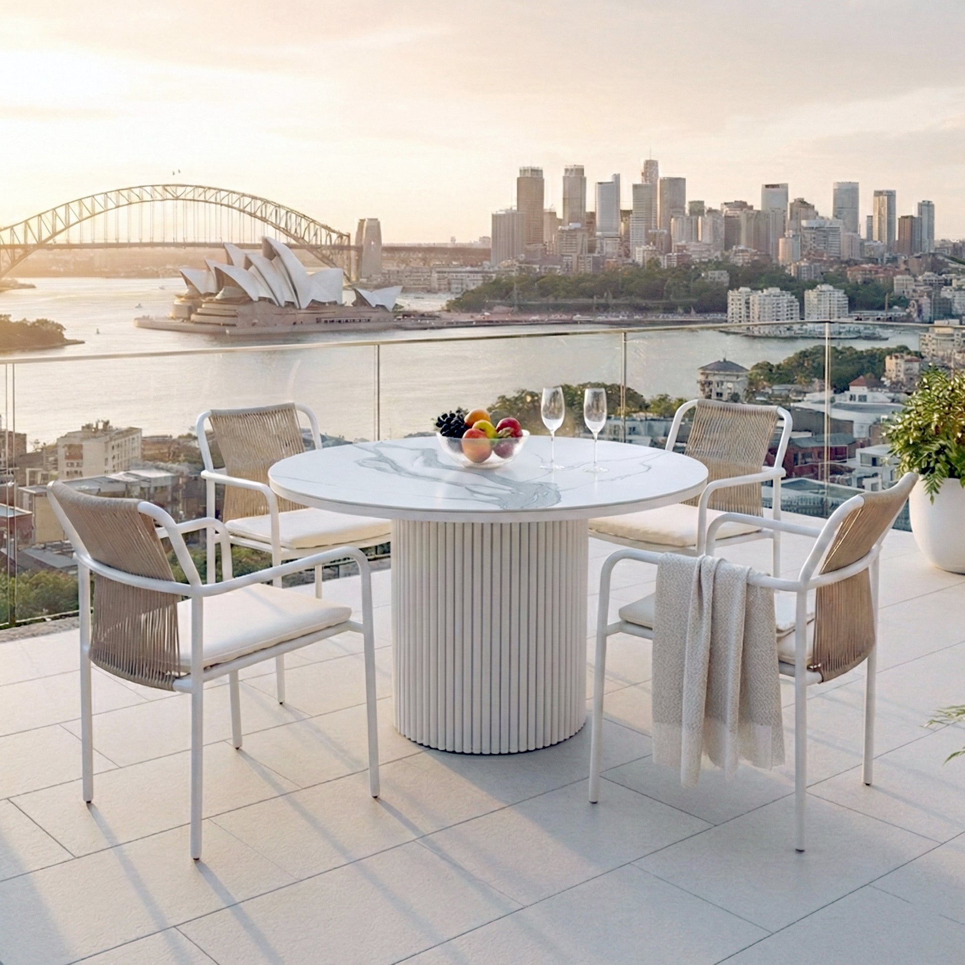 Evone 4 Seater Stone Outdoor Dining Table Set featuring a round marble top with four rope-back chairs, glass bowl of fruit, and wine glasses, overlooking a harbor at sunset.
