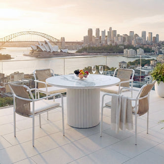 Evone 4 Seater Stone Outdoor Dining Table Set featuring a round marble top with four rope-back chairs, glass bowl of fruit, and wine glasses, overlooking a harbor at sunset.