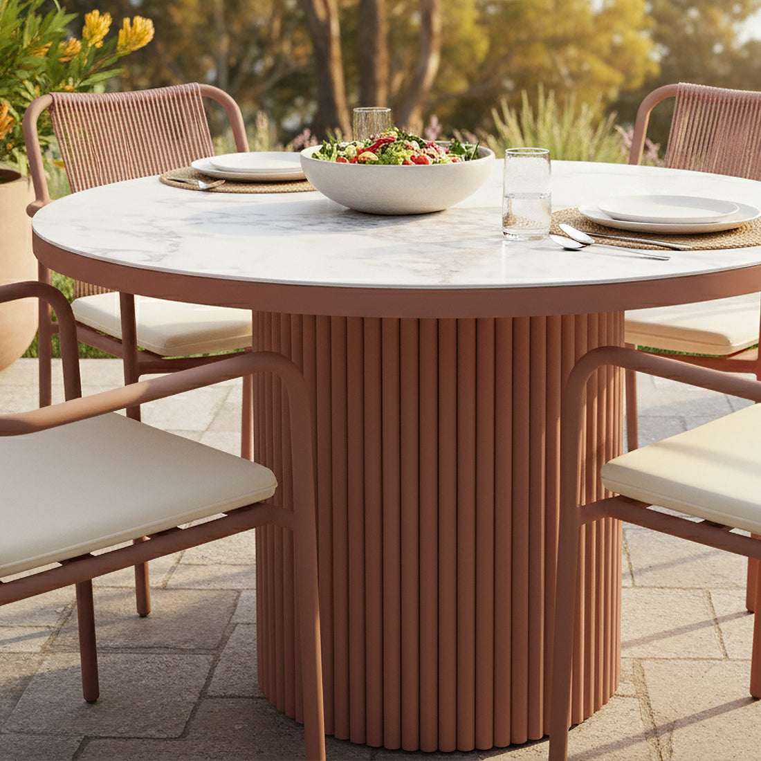 Evone Aluminium Outdoor Dining Table in Terracotta with a marble-like top, surrounded by matching chairs with light cushions and rope backs, set for a meal on a paved patio.